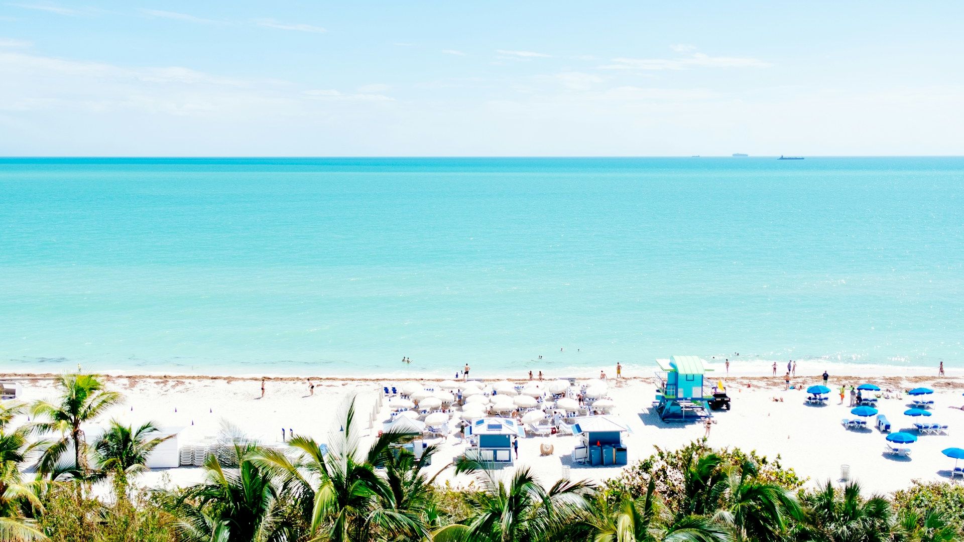 A pristine beach with white sand, clear turquoise water, and scattered beach amenities, surrounded by lush greenery under a clear sky.
