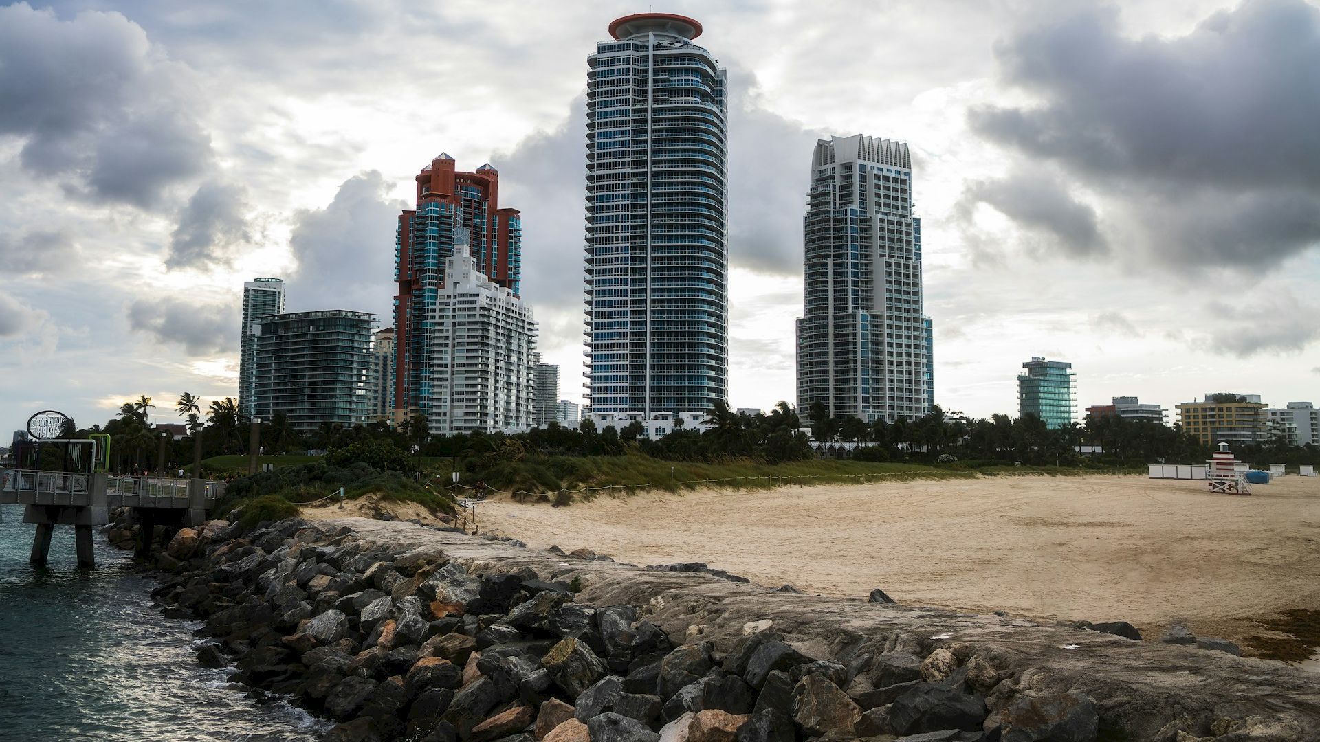 The image shows a rocky shoreline with a sandy area leading to a skyline of tall buildings under a partly cloudy sky.