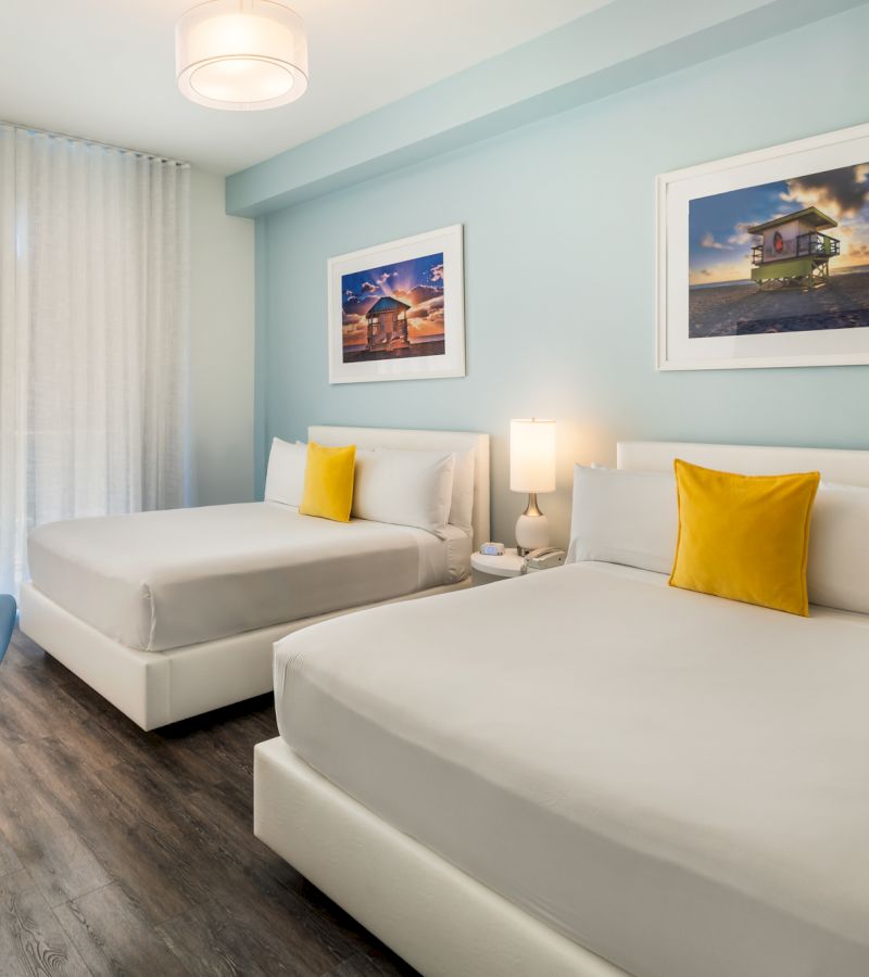 A hotel room with two beds, nightstand, lamp, and artwork on the walls.