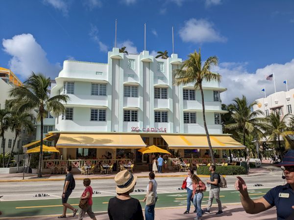 People walking on a sidewalk in front of a multi-story Art Deco building with yellow awnings, palm trees, and a clear blue sky.