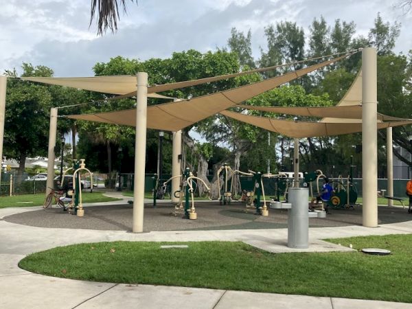 The image shows an outdoor fitness area with exercise equipment, covered by shade structures, surrounded by grass, trees, and a paved walkway.