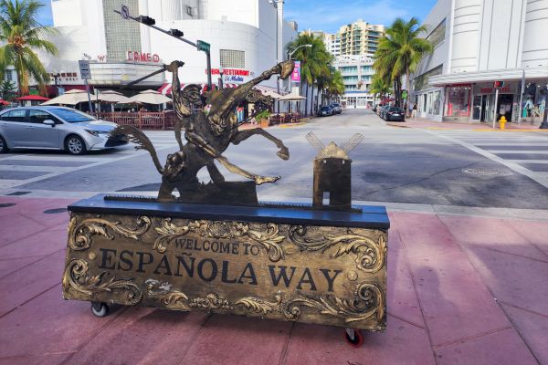 A decorative sign reads "Welcome to Española Way" on a street with palm trees, outdoor seating, and shops.