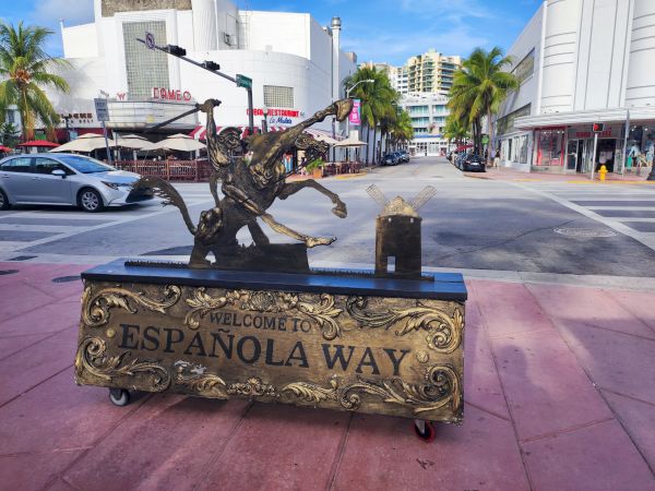 A decorative sign reads "Welcome to Española Way" on a street with palm trees, outdoor seating, and shops.