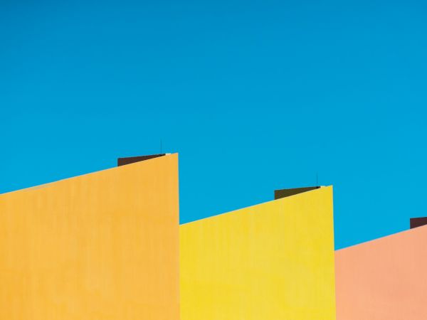 The image shows colorful, geometric building rooftops with vibrant shades of pink, orange, and yellow against a clear blue sky.