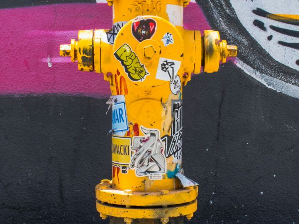 The image shows a bright yellow fire hydrant covered in various colorful stickers and graffiti, with a mural painted on the wall behind it.