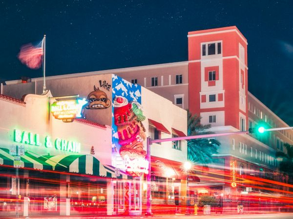 A vibrant night street scene with colorful buildings, neon lights, and light trails from passing vehicles. The sky is filled with stars.