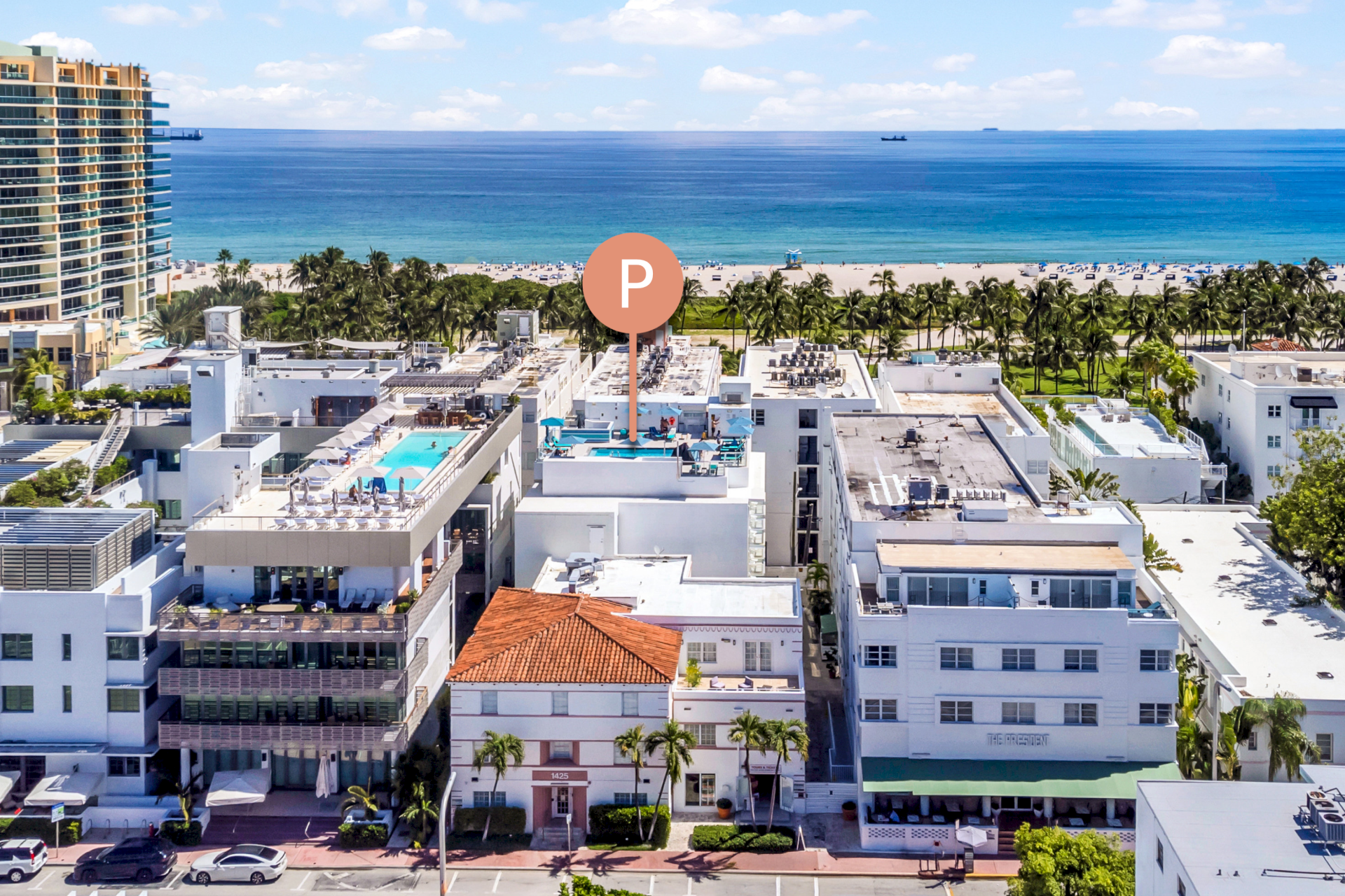 Aerial view of a white hotel building with a pool and palm trees, overlooking the beach and ocean.