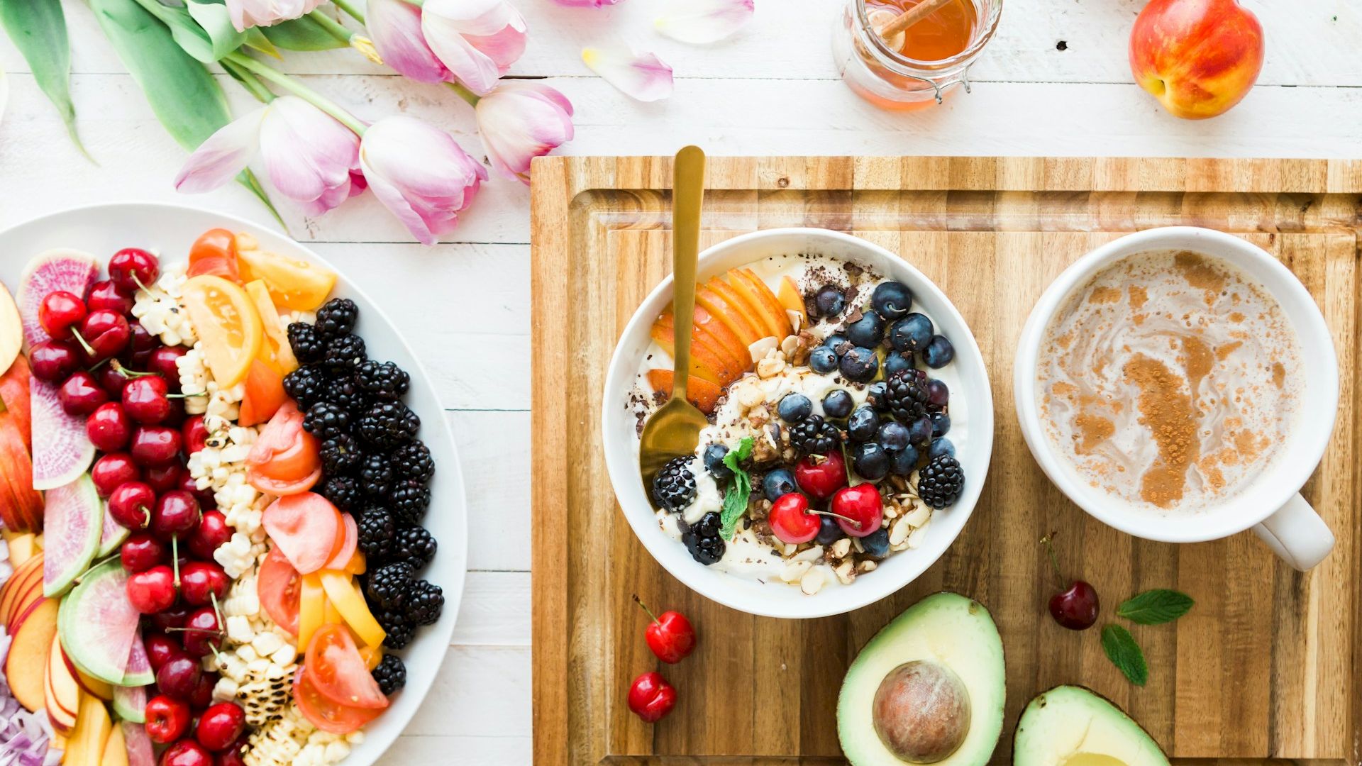 The image shows a healthy breakfast with bowls of yogurt and fresh fruits, a cup of coffee, an avocado, and some flowers on a white table.