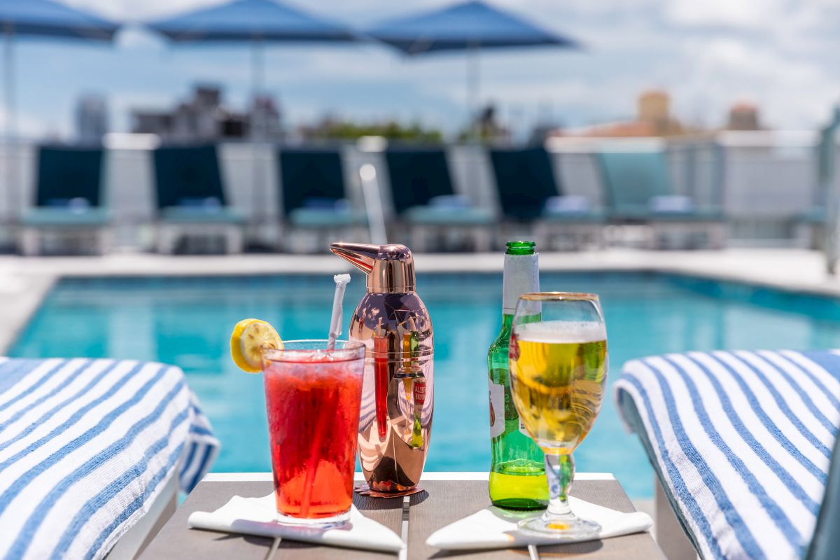 A table with drinks and towels in front of a pool with lounge chairs and umbrellas.