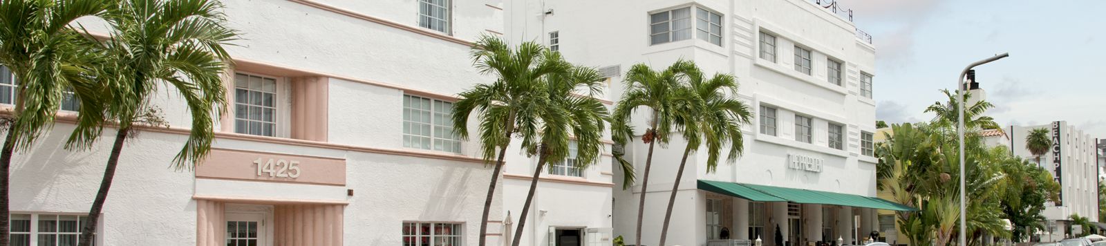 A sunny street with parked cars beside a white building and palm trees, with a “Beach” sign on the right.