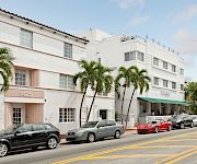 A sunny street with parked cars beside a white building and palm trees, with a “Beach” sign on the right.