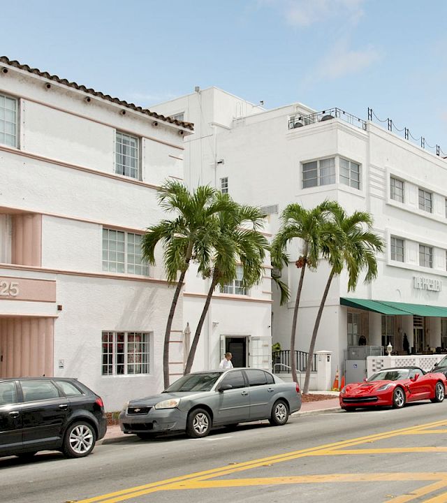 A sunny street with parked cars beside a white building and palm trees, with a “Beach” sign on the right.