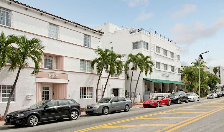 A sunny street with parked cars beside a white building and palm trees, with a “Beach” sign on the right.