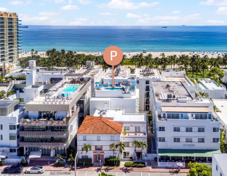 A bright oceanfront resort with palm trees, white buildings, and a sandy beach beside a calm blue sea under sunny clouds.