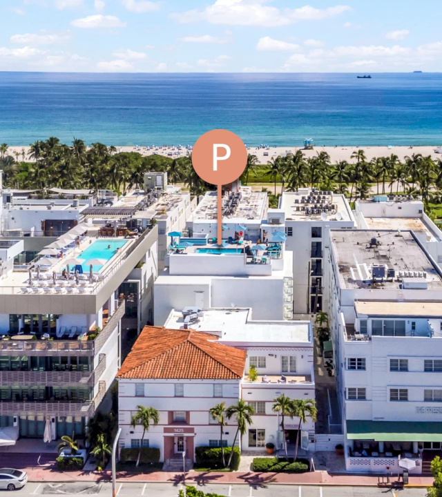 A bright oceanfront resort with palm trees, white buildings, and a sandy beach beside a calm blue sea under sunny clouds.
