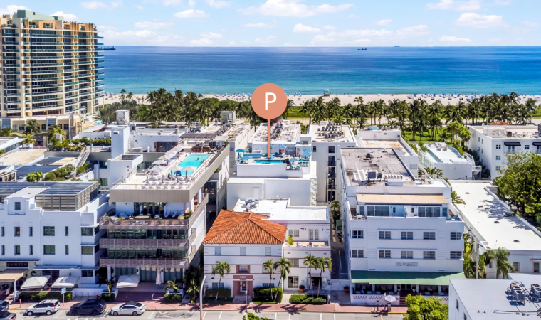 A bright oceanfront resort with palm trees, white buildings, and a sandy beach beside a calm blue sea under sunny clouds.