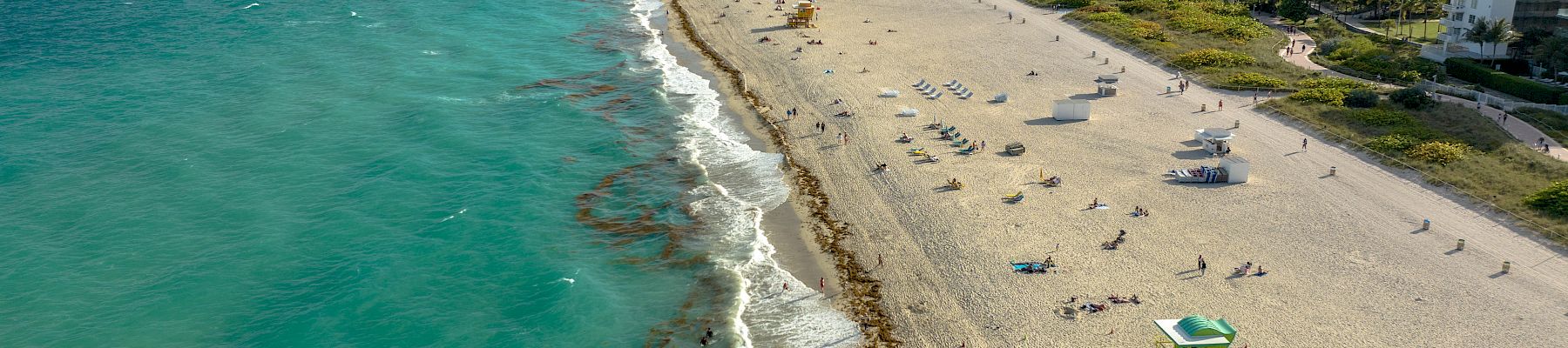 A sunny beachfront with turquoise ocean, sandy shore, and high-rise hotels/city skyline under dramatic clouds.