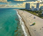 A sunny beachfront with turquoise ocean, sandy shore, and high-rise hotels/city skyline under dramatic clouds.