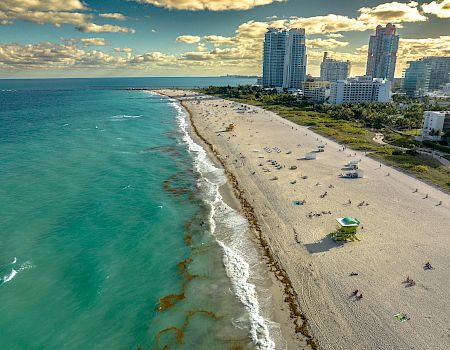 A sunny beachfront with turquoise ocean, sandy shore, and high-rise hotels/city skyline under dramatic clouds.