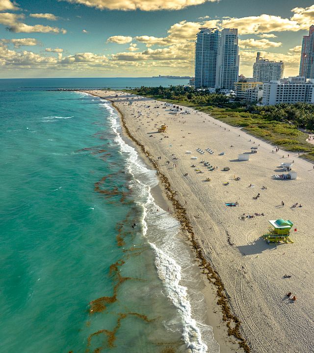 A sunny beachfront with turquoise ocean, sandy shore, and high-rise hotels/city skyline under dramatic clouds.