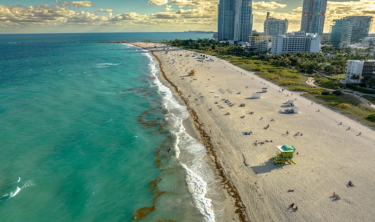 A sunny beachfront with turquoise ocean, sandy shore, and high-rise hotels/city skyline under dramatic clouds.