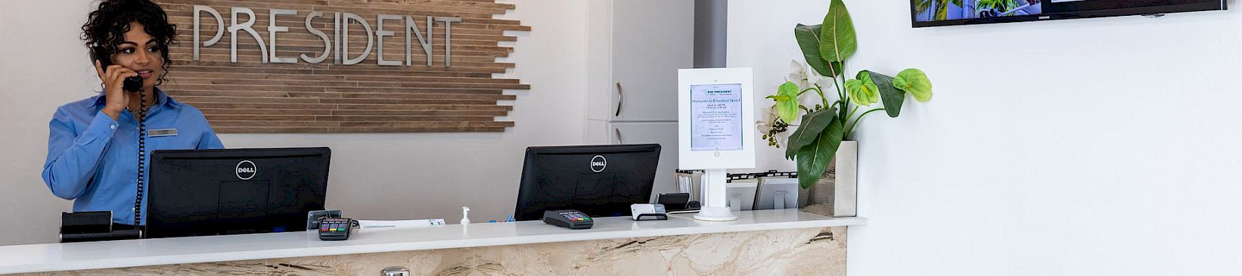 A hotel reception desk with a staff member on the phone, “President” sign behind, and a digital board reading Lincoln Road Mall.