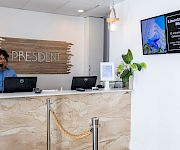 A hotel reception desk with a staff member on the phone, “President” sign behind, and a digital board reading Lincoln Road Mall.