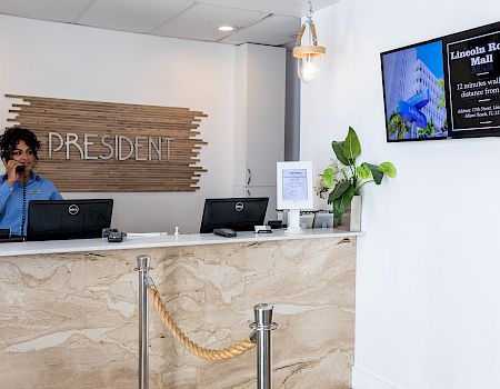 A hotel reception desk with a staff member on the phone, “President” sign behind, and a digital board reading Lincoln Road Mall.