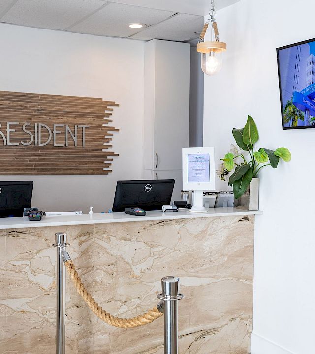 A hotel reception desk with a staff member on the phone, “President” sign behind, and a digital board reading Lincoln Road Mall.