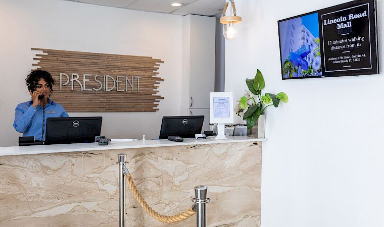 A hotel reception desk with a staff member on the phone, “President” sign behind, and a digital board reading Lincoln Road Mall.