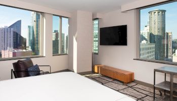 A modern hotel room with large windows showcasing a cityscape, a bed, a TV, a chair, and a small table.