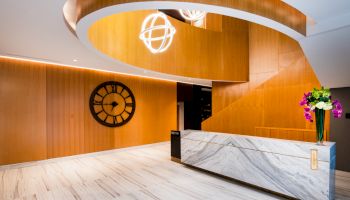 The image shows a modern lobby with a white marble reception desk, a large wall clock, a winding staircase, and decorative ceiling lights.