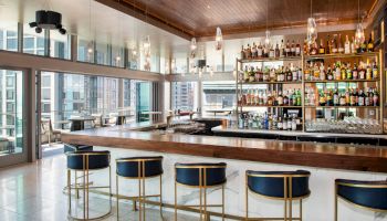 A modern bar with sleek stools, a well-stocked liquor shelf, and large windows overlooking a cityscape, creating a stylish atmosphere.
