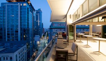 The image shows a modern cityscape with tall buildings at dusk, and an elegant rooftop patio featuring tables and chairs.