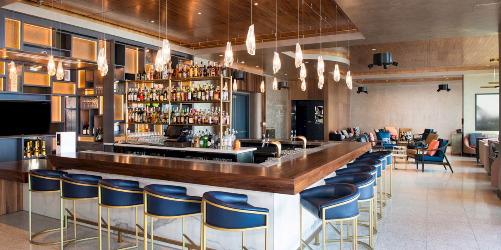 Modern bar with a wooden counter, blue chairs, pendant lights, and well-stocked shelves. Cozy seating area in the background.