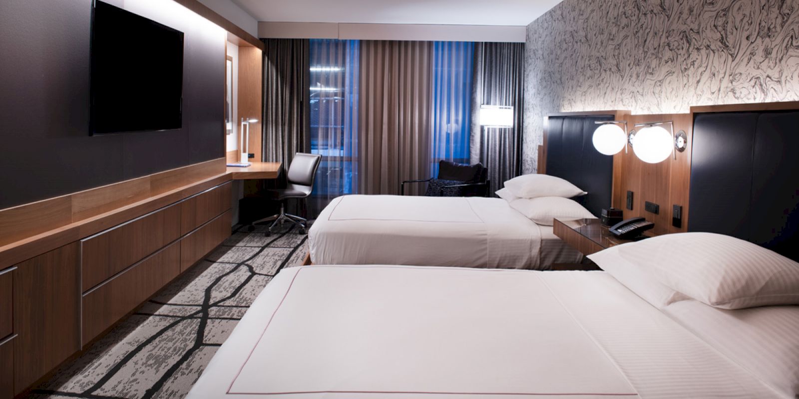 A modern hotel room with two double beds, a flat-screen TV, a desk with a chair, and patterned carpet, all in neutral tones.