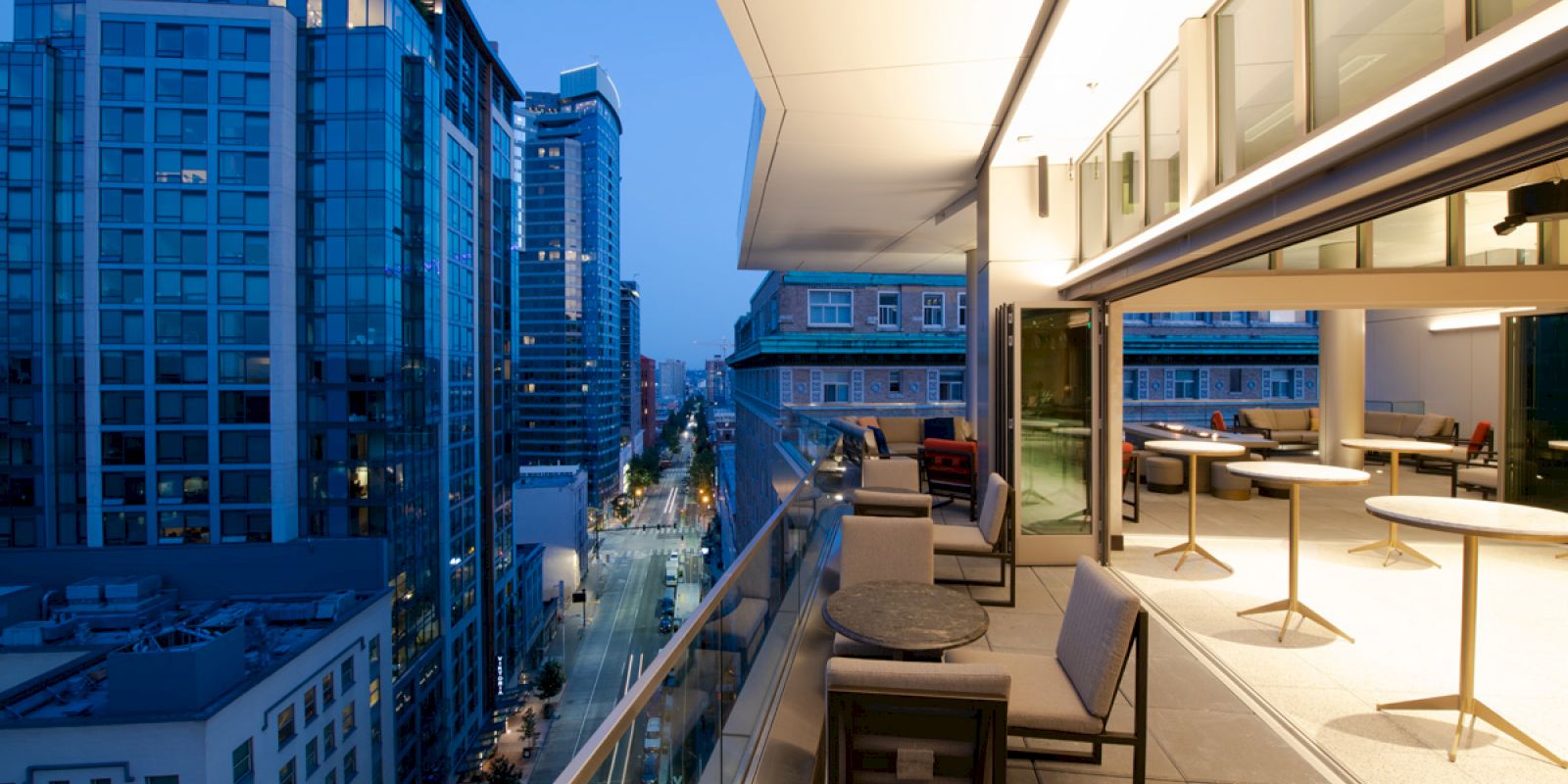 A modern balcony with seating overlooks an urban street scene at dusk, lined with glassy high-rise buildings and lights.