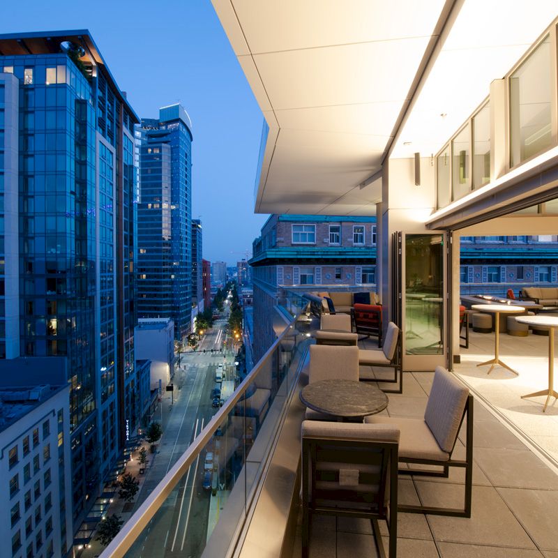 An outdoor terrace with seating overlooks a cityscape at dusk, featuring modern buildings and an adjacent open indoor area with tables.