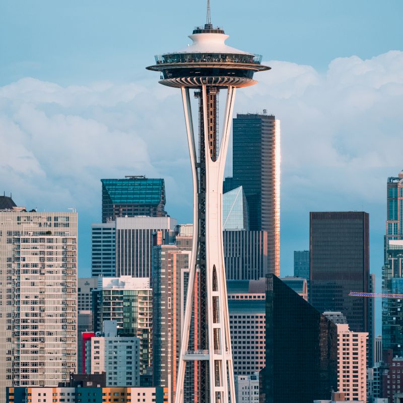 The image shows the iconic Space Needle with Seattle's skyscrapers in the background, under a cloudy sky and urban landscape.