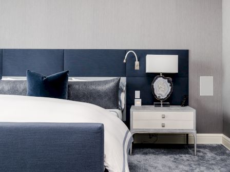 A modern bedroom with a blue bed, gray and white pillows, a white nightstand with a lamp, and a decorative item on a plush carpeted floor.