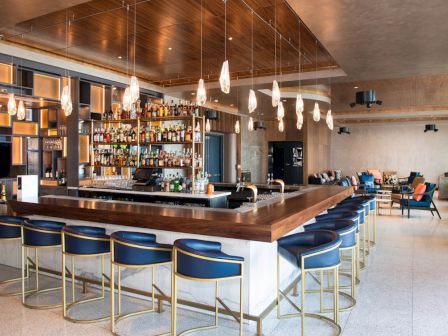 A modern bar with a wooden counter, blue chairs, hanging lights, and a well-stocked back wall. The room has a cozy seating area.
