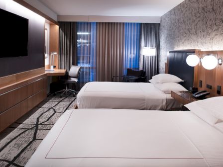 A modern hotel room with two beds, a TV, desk, and chairs. The room has stylish decor and lighting, with patterned walls and carpeting.