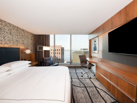 A modern hotel room with a large bed, flat-screen TV, desk, and a window view of city buildings. Neutral tones and contemporary decor.