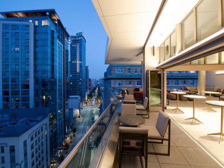 A modern cityscape at dusk with tall buildings and a stylish, open balcony featuring tables and chairs overlooking the street.