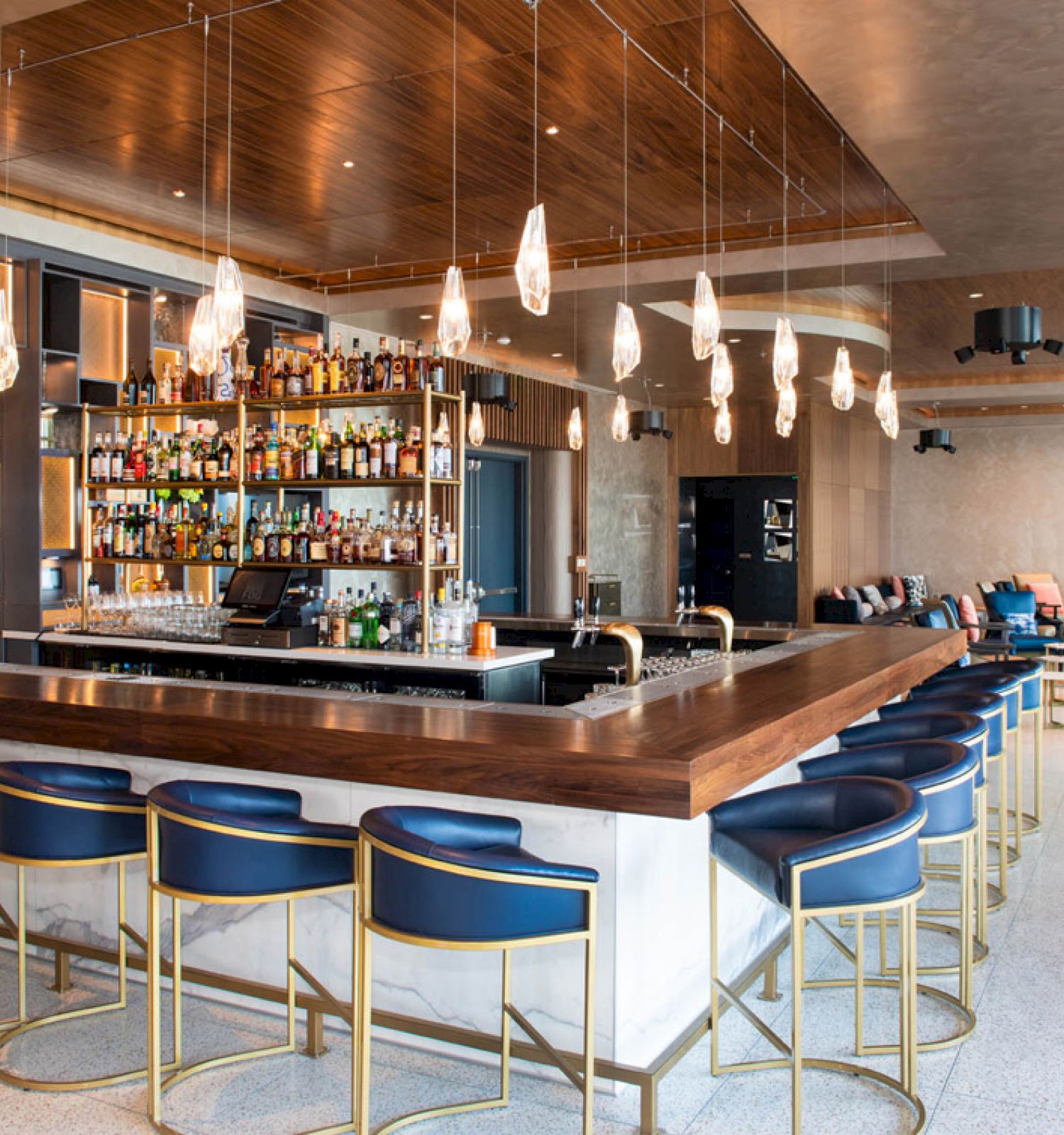 A modern bar with a stocked shelf, stylish seating, hanging lights, and a cozy lounge area in an open, contemporary setting.