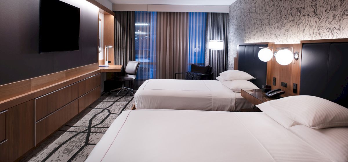 A modern hotel room with two beds, a wall-mounted TV, a desk with a chair, and warm lighting creates a cozy ambiance.