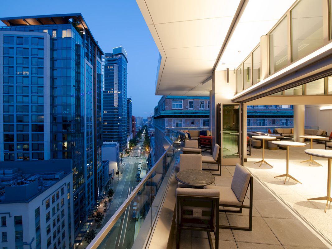 A modern balcony with furniture overlooks a cityscape featuring tall buildings and well-lit streets during twilight.