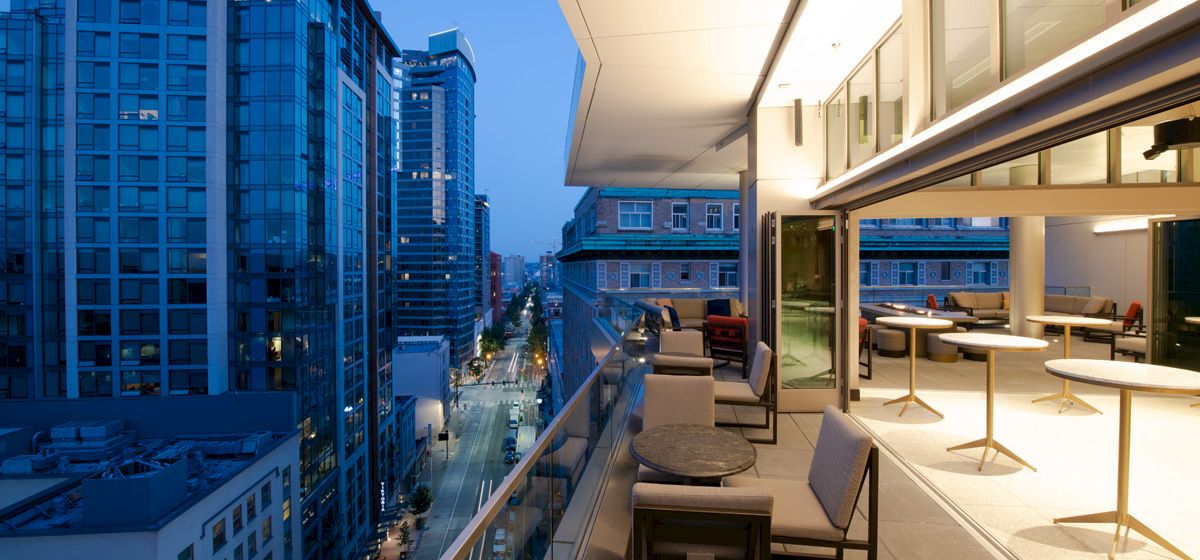 A modern balcony with furniture overlooks a cityscape featuring tall buildings and well-lit streets during twilight.