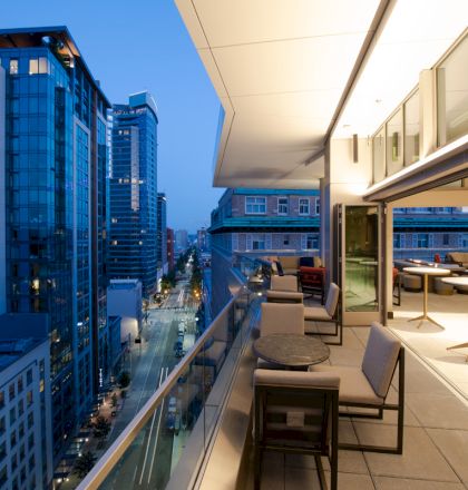 A modern cityscape with tall buildings and an elegant balcony area, featuring chairs and tables, at dusk.