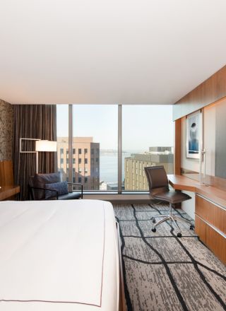 A modern hotel room with a large bed, desk, chair, and TV. The window offers a view of city buildings and a body of water.